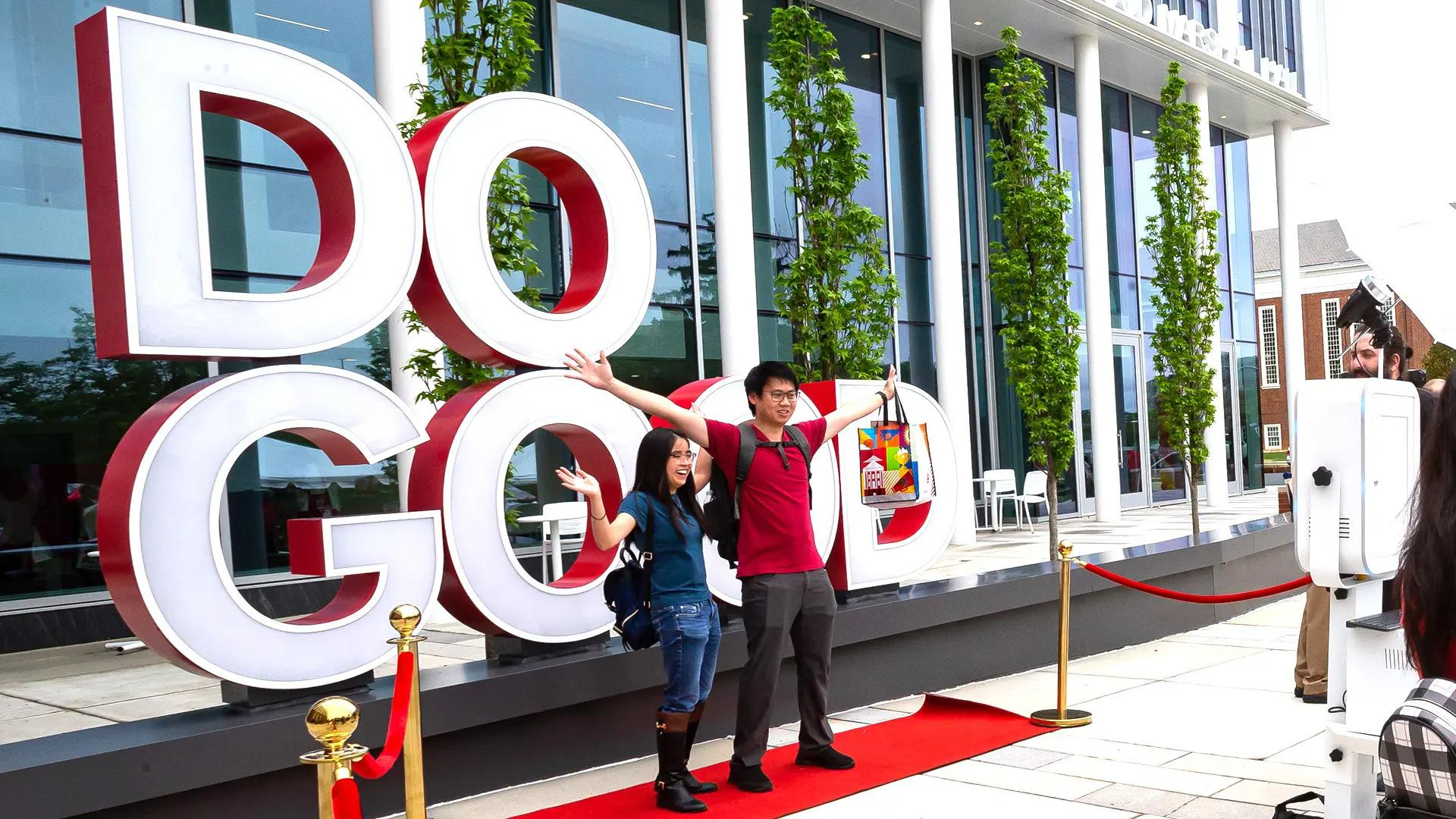 A man and a woman are standing with arms raised in front of a large Do Good sign. 