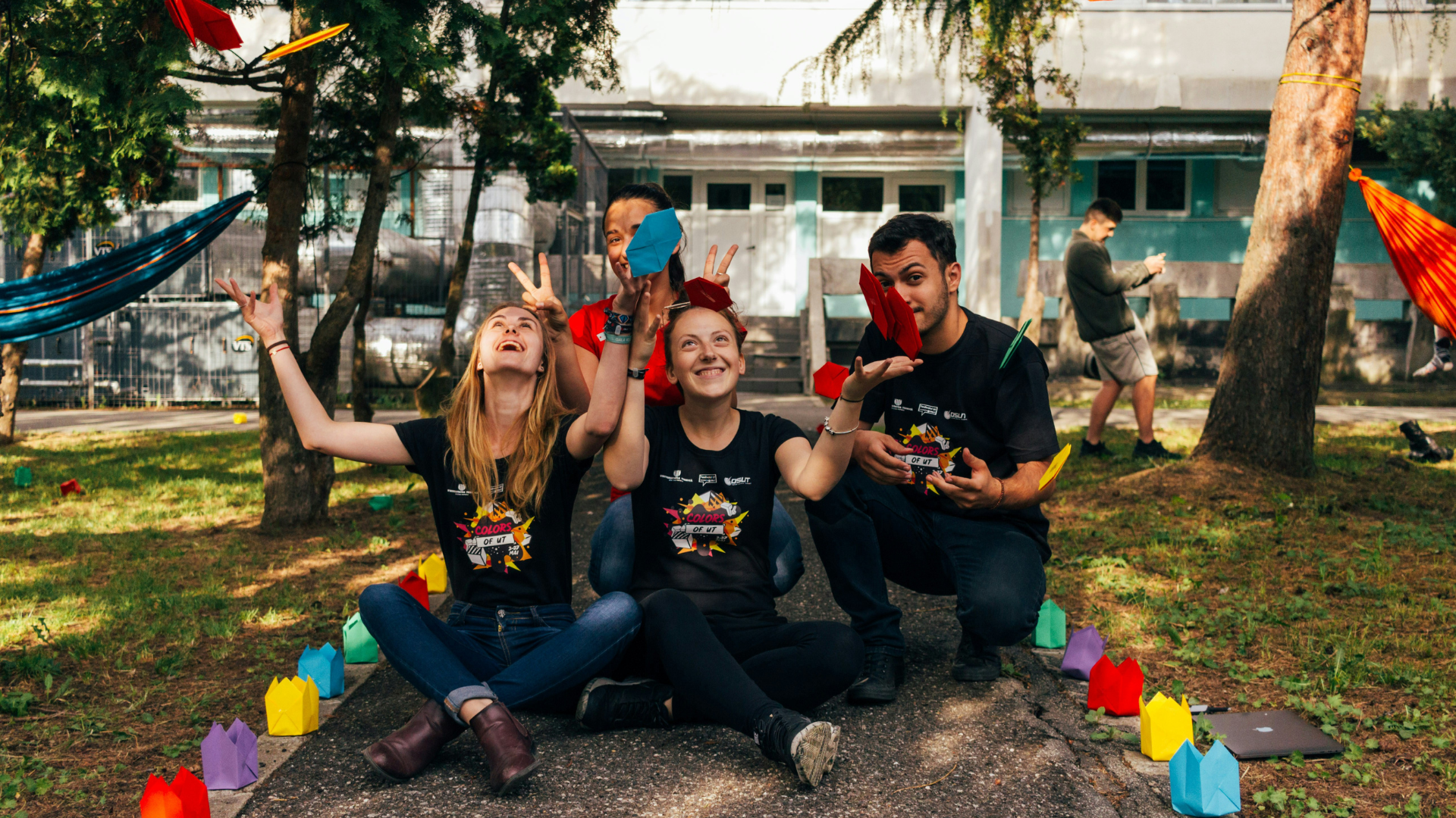 A group of three volunteers sitting down cross legged. Wearing black tshirts, they are throwing colorful pieces of fabric in the air. 