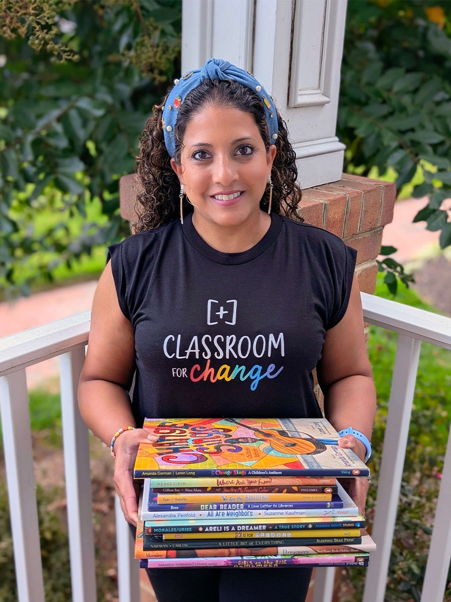 Rose Cherussery stands on a balcony with green trees and grass behind her holding a color stack of books in a black shirt with black hair