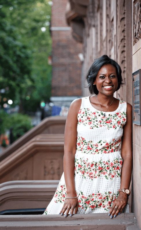 Ebonie is smiling as she leans up against a brick wall with trees in the background. 