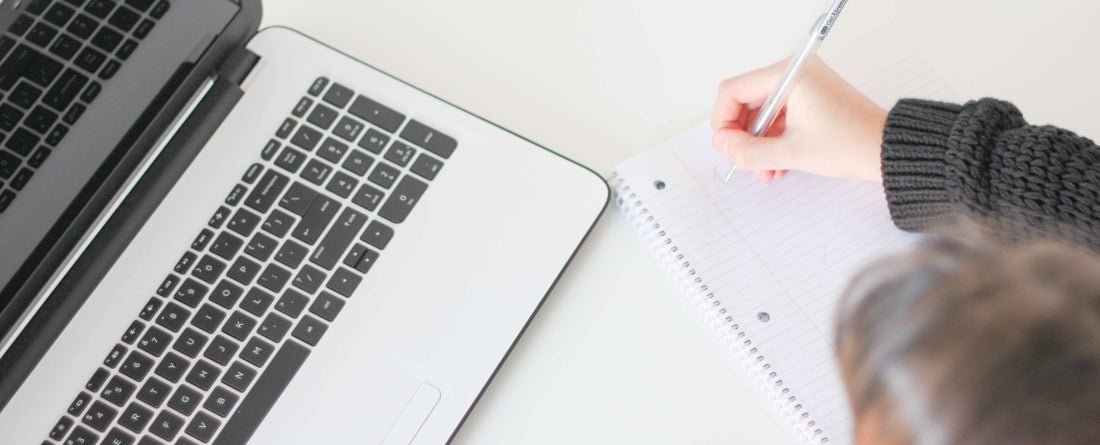 Person writing on notebook in front of laptop