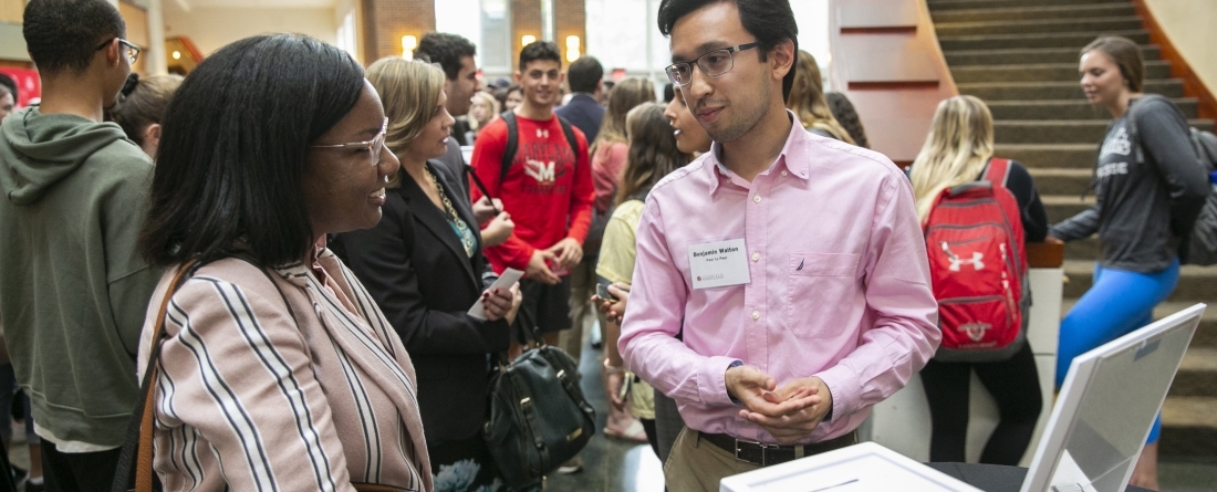 A male student is giving a brief pitch to a woman who is smiling and listening as he talks. 