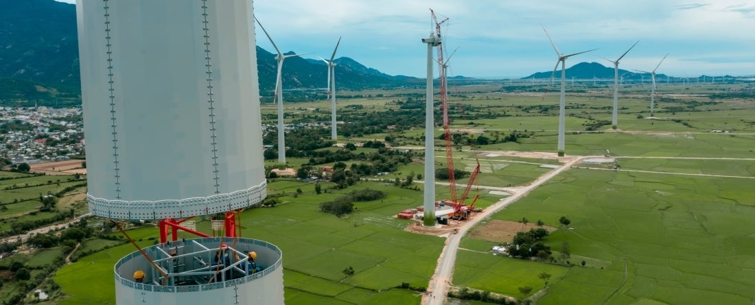 Wind turbines are under construction in green fields with a mountainous background on a day with blue skies.