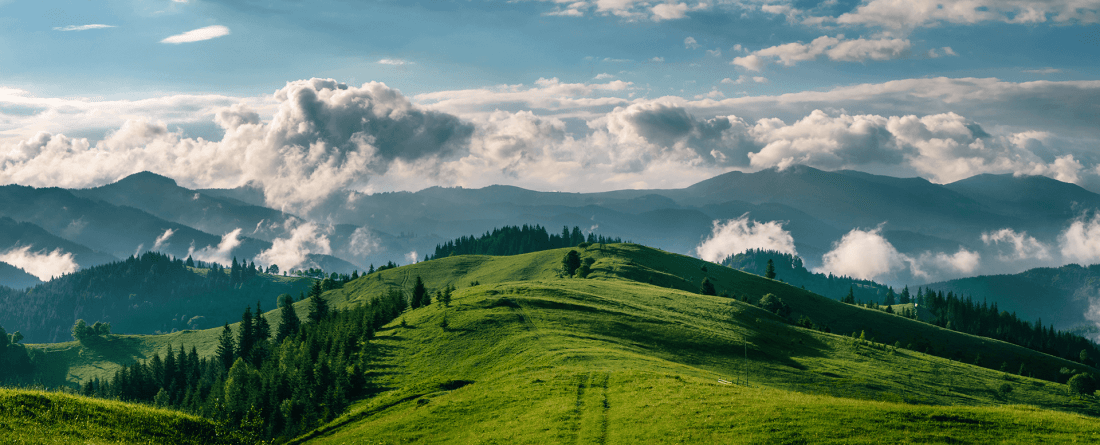 A grassy mountainous area with blue skies and clouds that rest above mountains in the background.
