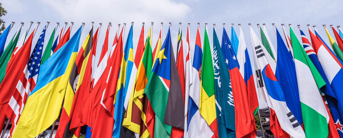 A set of international flags are placed next to each other in a row on a day with blue skies and clouds.