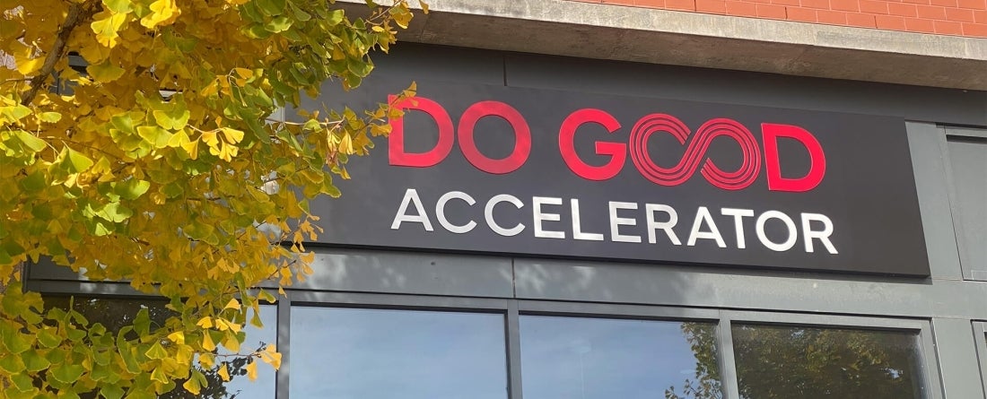 A yellow and green tree stands in front of a black metal sign that reads Do Good in red and Accelerator in white