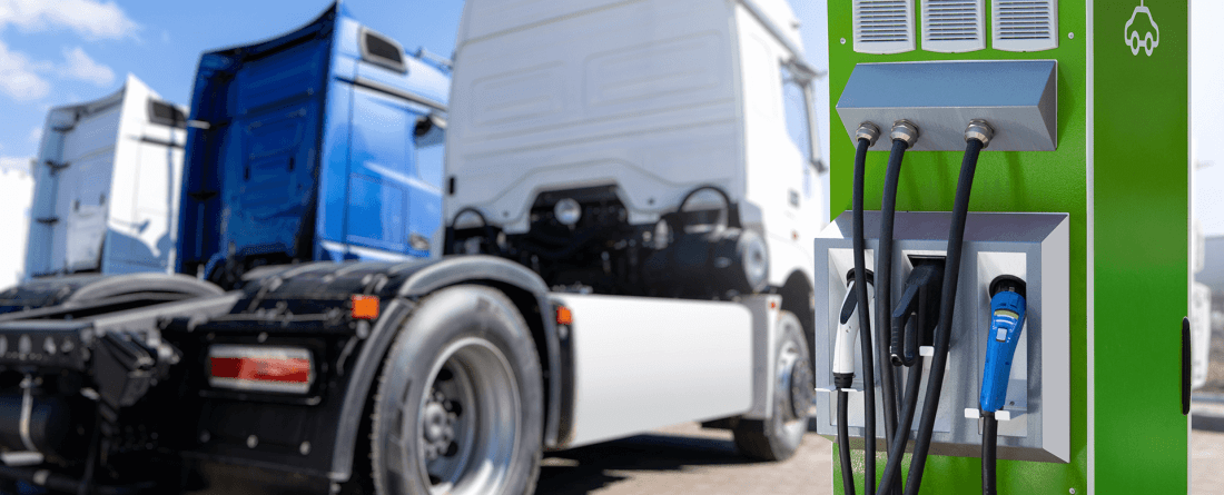 A green utility box with electric vehicle charging ports. Large heavy-duty vehicles are parked in the background.