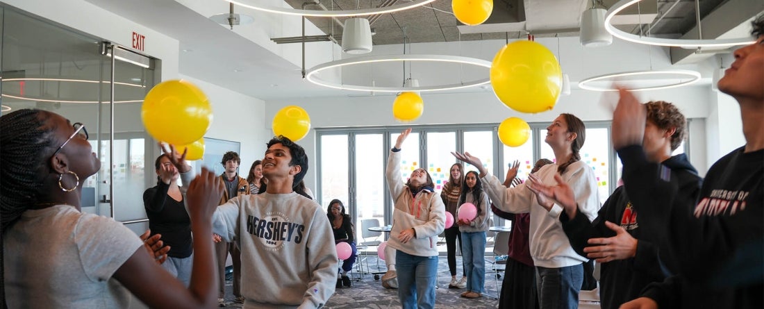 Students in a bright room with tall windows keep a yellow balloon afloat in the air