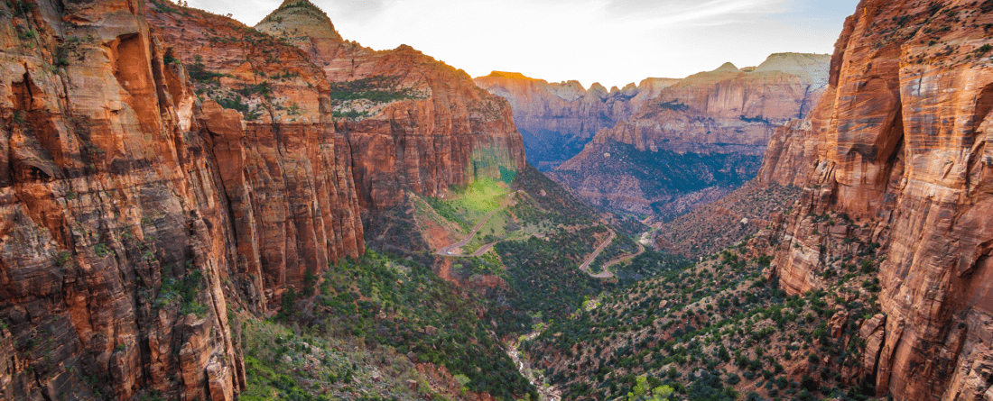 A canyon with orange rocks on a day with blue skies and sun. Green trees and other shrubs sit alongside a river at the bottom of the canyon.