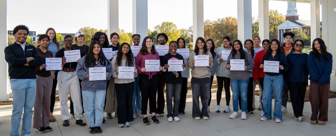 A large group of students and mentors pose for a photo on a rooftop with bright blue clouds in the background and tall white pillars.