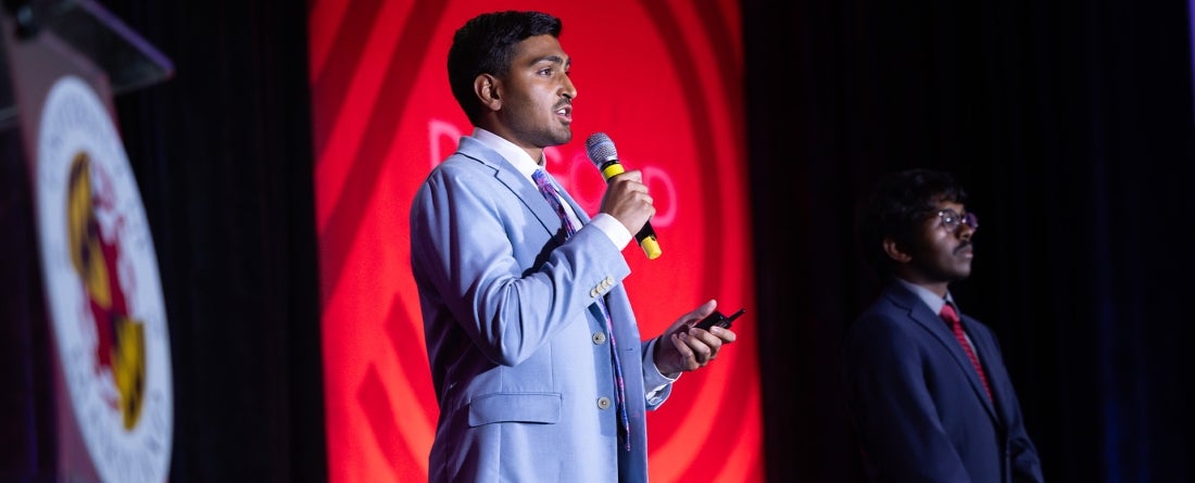 Two college students speaking on a stage with a red banner in the background.