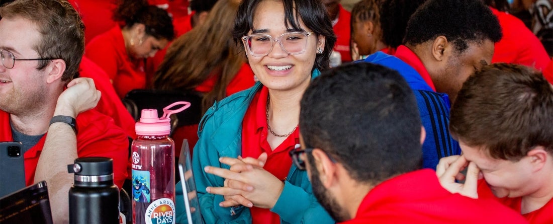Image is of a young woman with glasses smiling at a person she's in conversation with. There are other people behind her wearing red and talking.