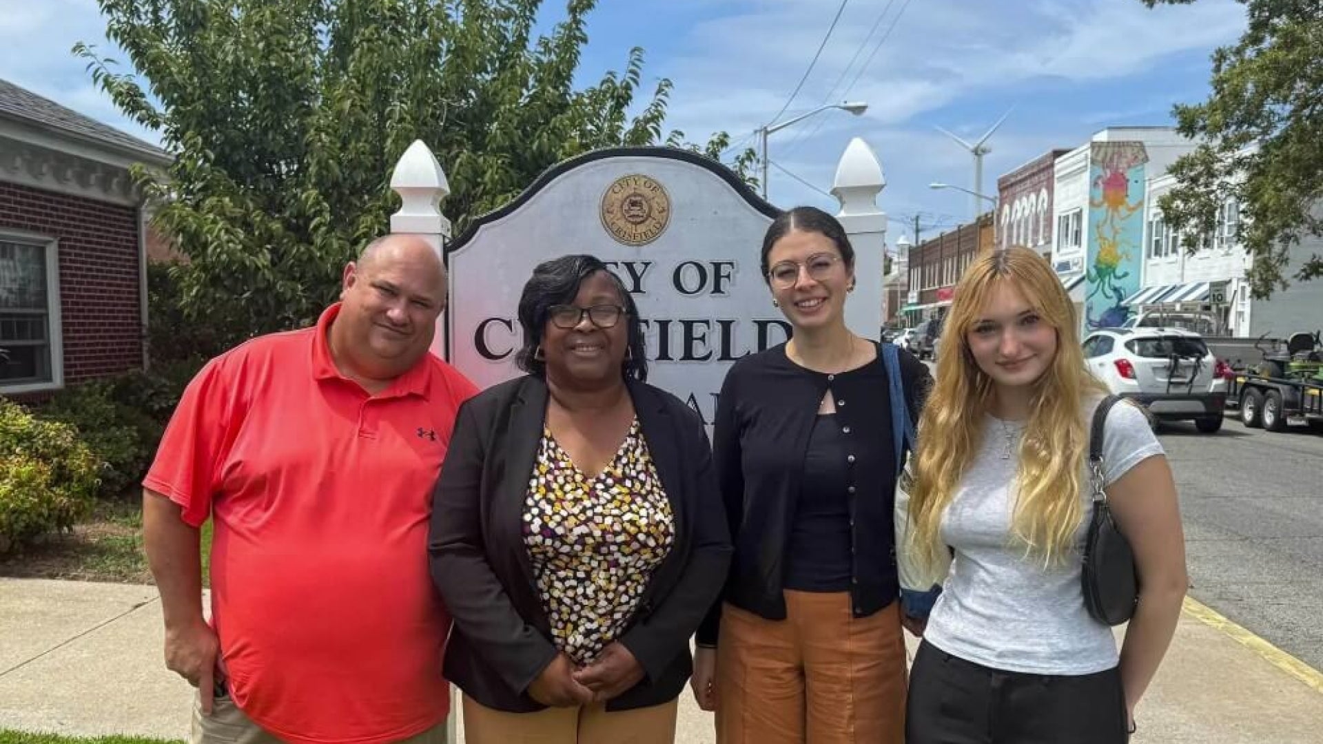 1 man and 3 women stand in front of a sign with a brick house to the left in front of a city sign and a tree on an urban street.